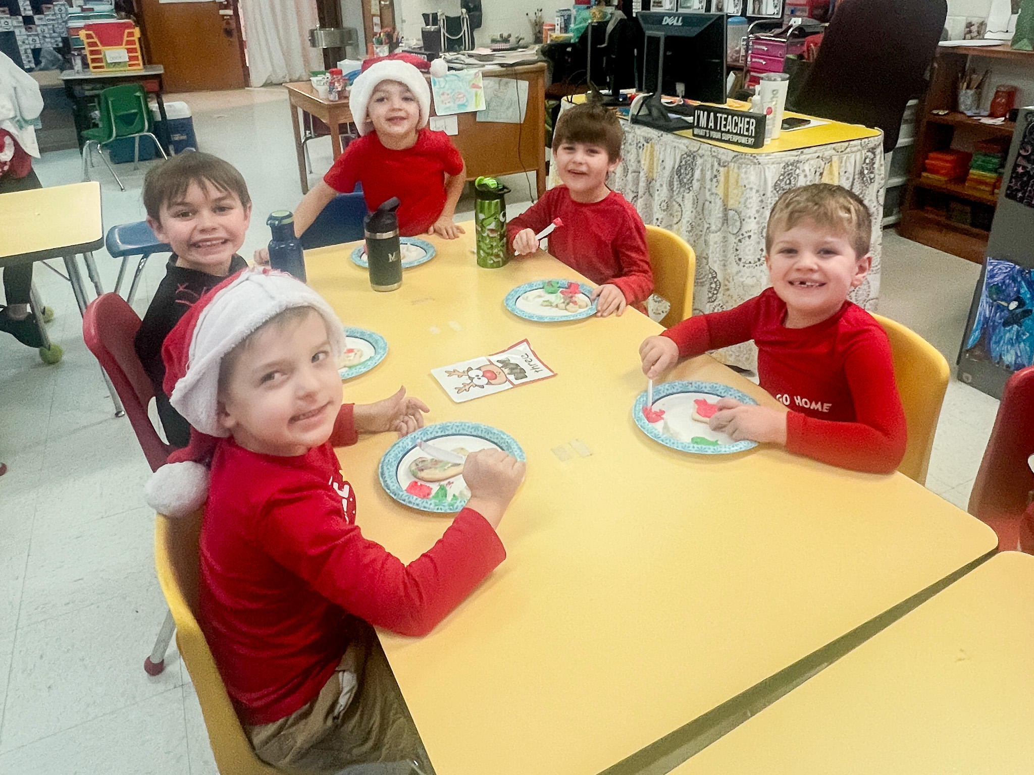 Five young boys sit at a table wearing red while decorating cookies